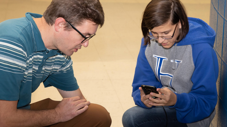 Two people sitting cross-legged on the floor, looking at a cell phone